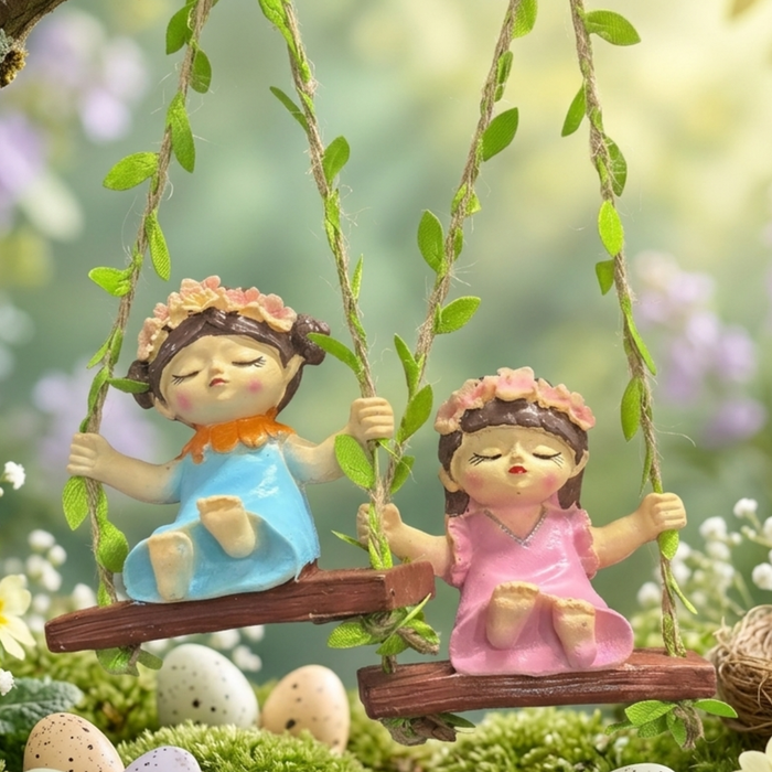 Two whimsical hand-painted girl figurines wearing flower crowns and colorful dresses (pink and blue), sitting on wooden-style swings decorated with green vines and hanging from twine.