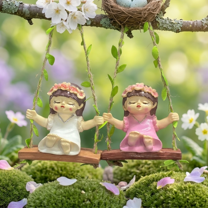 A whimsical resin figurine of a small girl with a pink flower crown and white dress sitting on a wooden swing suspended by twine ropes with green leaf accents.