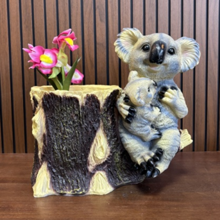 A decorative resin planter shaped like a tree stump featuring a mother koala holding her joey, with pink artificial flowers placed inside the planting well.