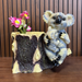 A decorative resin planter shaped like a tree stump featuring a mother koala holding her joey, with pink artificial flowers placed inside the planting well.