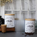 Two white metal storage canisters labeled "Homemade Tradition Tea" and "Homemade Tradition Coffee" with wooden lids, displayed on a kitchen counter with a glass of tea and coffee beans.