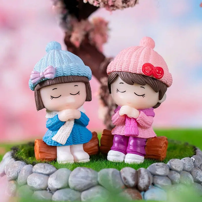 Two small figurines of a girl and a boy wearing knitted hats and scarves, sitting on rocks with a blurred floral background.