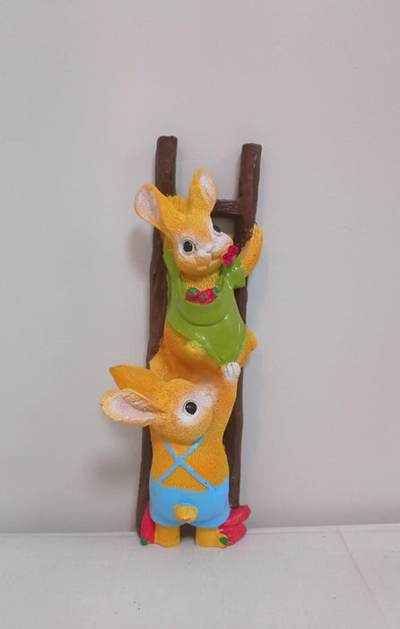 A colorful garden or shelf figurine depicting two yellow rabbits climbing a brown ladder. The top rabbit wears a green apron and is holding a red flower, while the bottom rabbit wears blue overalls and is helping the top one climb.