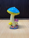 A whimsical trio mushroom garden statue featuring a large blue cap, a medium pink cap, and a small yellow cap, all with white spots and realistic textured stems on a rocky base with tiny red flowers.