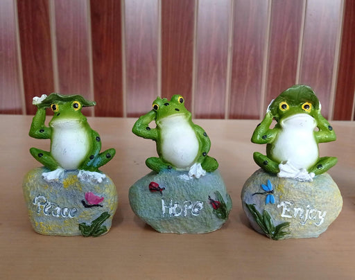 Three resin frog figurines, each sitting on a painted rock. They are posed similarly to the "Three Wise Monkeys" (see no evil, hear no evil, speak no evil), but as frogs. The rocks are labeled "Peace," "Hope," and "Enjoy" in cursive lettering, with painted details like butterflies, ladybugs, and dragonflies.