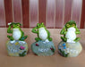 Three resin frog figurines, each sitting on a painted rock. They are posed similarly to the "Three Wise Monkeys" (see no evil, hear no evil, speak no evil), but as frogs. The rocks are labeled "Peace," "Hope," and "Enjoy" in cursive lettering, with painted details like butterflies, ladybugs, and dragonflies.