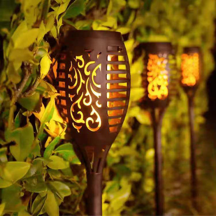 A set of four solar-powered flickering flame torches illuminating a garden path near a white fence.