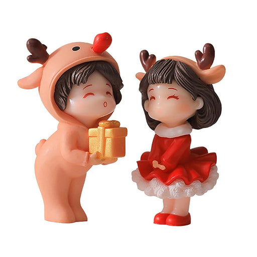 Two miniature Christmas figures. A boy figure in a tan reindeer costume is holding a gold gift box towards a girl figure wearing a red Santa-style dress and a reindeer headband.