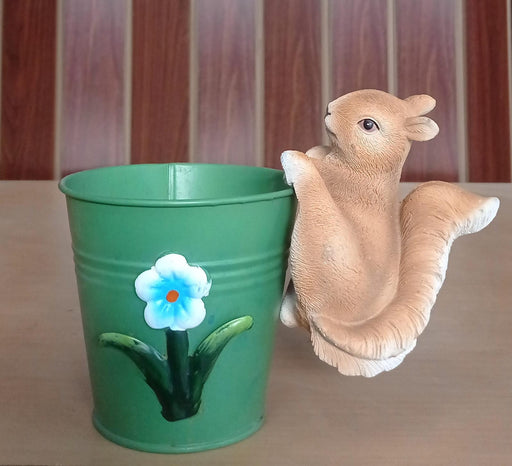 A close-up of a small, realistic resin or ceramic squirrel figurine, colored in natural light brown and white, clinging to the side of a  pot.