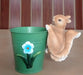 A close-up of a small, realistic resin or ceramic squirrel figurine, colored in natural light brown and white, clinging to the side of a  pot.