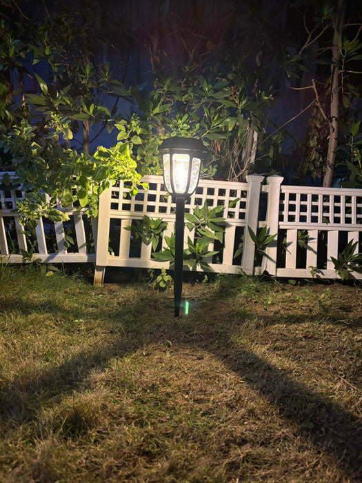 "Black solar-powered garden pathway light glowing at night in front of a white lattice fence and green bushes."