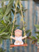 A whimsical garden figurine of a small girl with a flower crown sitting on a wooden swing. The swing is hung from a tree trunk by jute ropes entwined with artificial green leaves, set against a background of a white picket fence and greenery.