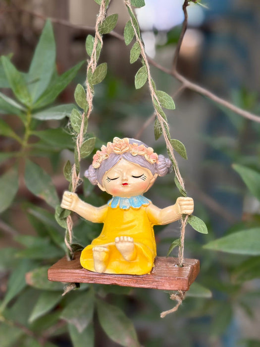A whimsical garden figurine of a young girl with a floral crown and yellow dress sitting on a wooden swing suspended by leaf-entwined twine.