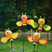 Four cute, smiling honey bee garden stakes with bright orange and yellow bodies and colorful wings, standing in a garden with a blurred fence background.
