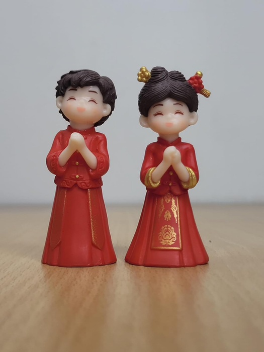A pair of miniature figurines representing a Chinese wedding couple in traditional red and gold clothing, standing with hands pressed together in a greeting on a wooden surface.