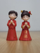 A pair of miniature figurines representing a Chinese wedding couple in traditional red and gold clothing, standing with hands pressed together in a greeting on a wooden surface.