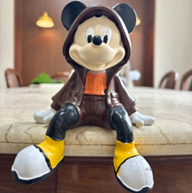 A collectible Mickey Mouse figurine wearing a brown hooded jacket, orange shirt, and yellow and white boots, sitting on a marble surface.