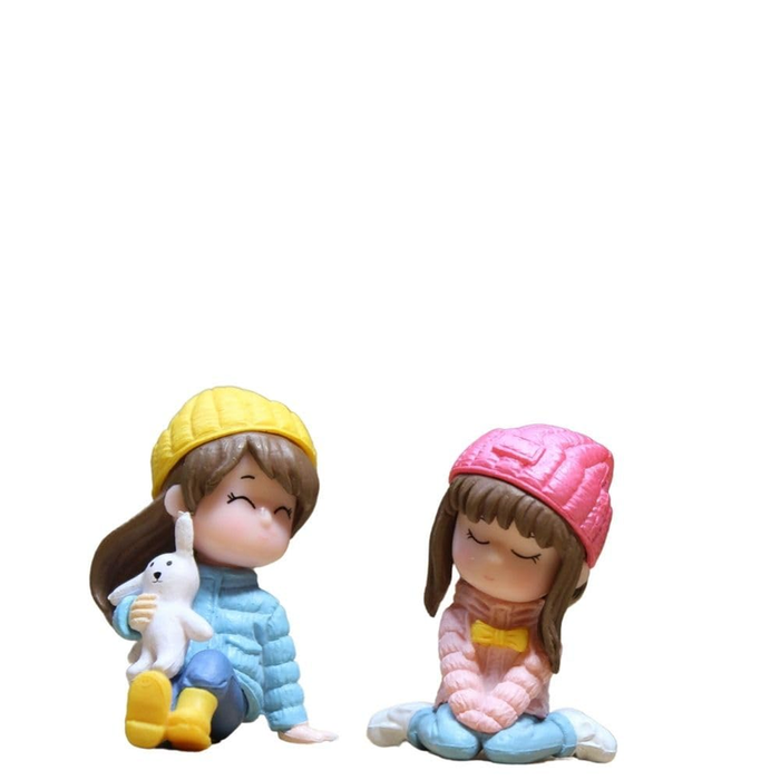Two miniature figurines of girls in winter gear; one wearing a yellow beanie and blue jacket holding a white rabbit, and another in a pink beanie and pink jacket sitting cross-legged.