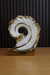 A modern ceramic sculpture of a swirling white feather with shiny gold-plated edges, standing on a rectangular gold base against a dark slatted background.