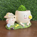 Two cute, smiling egg-shaped character planters sitting on a mossy base; the larger egg holds a yellow flower while the smaller one wears a sun hat and holds a miniature shovel.