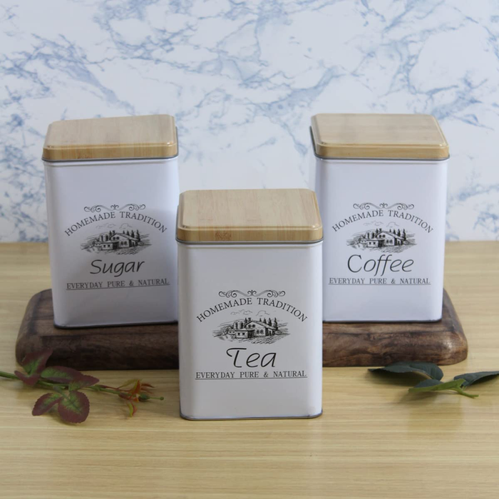 Three white rectangular metal storage canisters labeled Sugar, Tea, and Coffee with wooden lids, arranged on a wooden tray