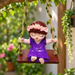 Cute resin girl figurine in a purple dress and flower crown hanging on an ivy-wrapped swing outdoors.