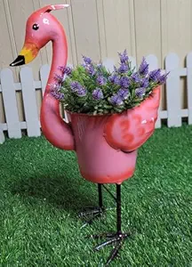 A bright pink metal flamingo planter filled with faux purple lavender flowers, standing on artificial grass in front of a white picket fence.