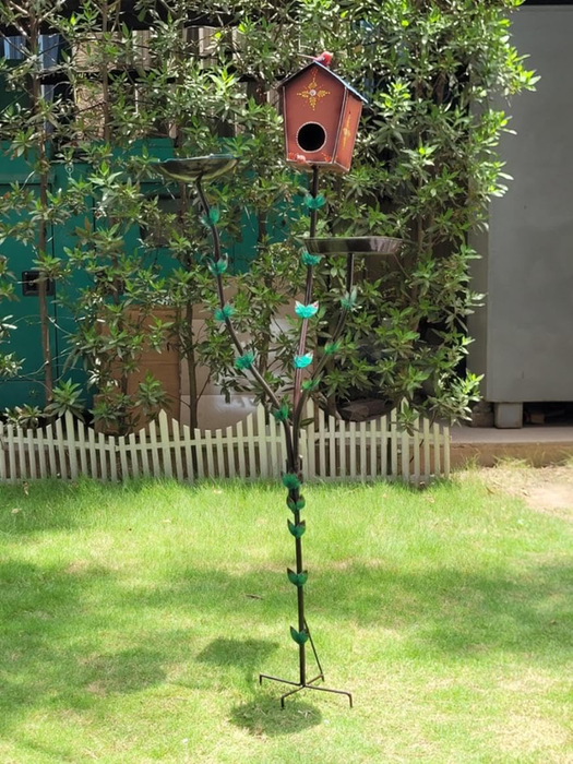 A tall metal garden stand shaped like a vine with green leaves, featuring a brown birdhouse on top and two metal feeder trays, standing on a green lawn.