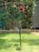 A tall metal garden stand shaped like a vine with green leaves, featuring a brown birdhouse on top and two metal feeder trays, standing on a green lawn.