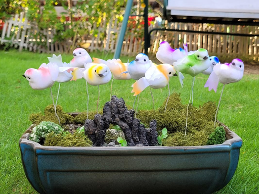 A collection of colorful miniature artificial birds on wire stems, arranged in a ceramic planter with moss and a small stone archway.