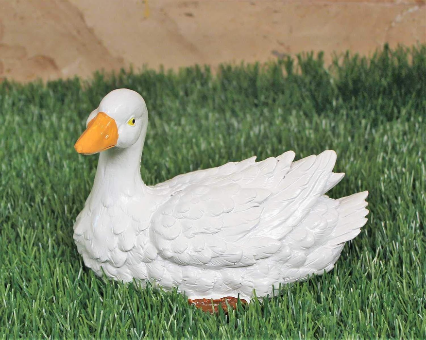 A miniature white goose figurine with an orange beak resting on green artificial grass against a stone-textured background.