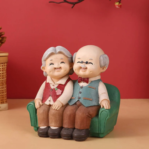 A whimsical handcrafted figurine of a smiling elderly couple sitting closely together on a green sofa, featuring a grandmother in a red vest and a grandfather in a teal vest with a red bow tie