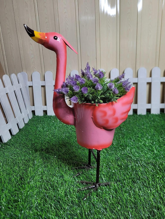 A side view of a standing pink metal flamingo planter filled with faux purple flowers, placed on green artificial turf with a white fence in the background.