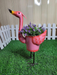 A side view of a standing pink metal flamingo planter filled with faux purple flowers, placed on green artificial turf with a white fence in the background.
