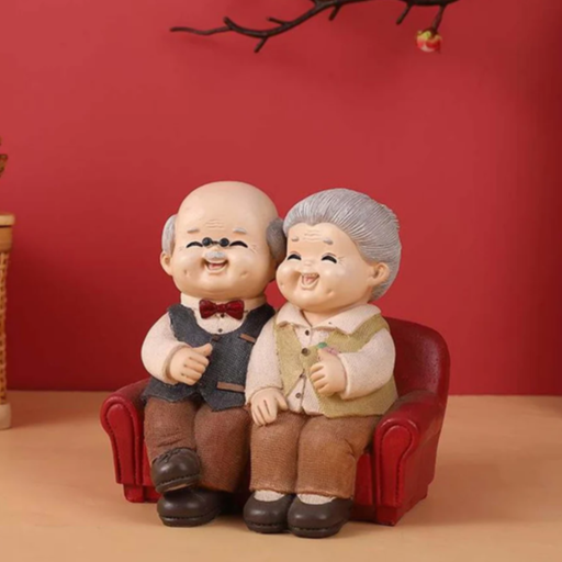 A handcrafted resin figurine of a smiling elderly couple sitting closely together on a miniature red sofa, featuring a grandfather in a vest and bowtie and a grandmother with grey hair, set against a red background.