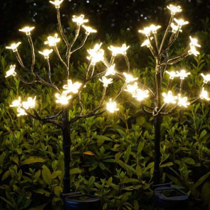 Two solar-powered LED blossom tree garden stakes glowing with warm white lights nestled among green foliage at dusk