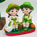 Hand-painted figurine of a young boy and girl in green garden outfits sitting on a log with a white duck and a small orange pumpkin.