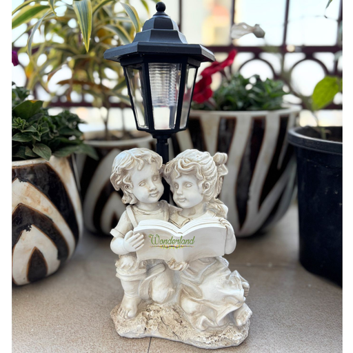 White resin garden statue of a boy and girl reading a book under a solar-powered black vintage lamp post on a grass lawn.