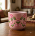 Handpainted pink terracotta flower pot featuring an intricate traditional lotus flower and leaf design resting on a wooden table.
