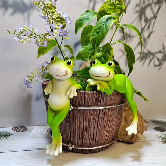 Two cute hand-painted resin frog figurines hanging over the rim of a flower pot filled with green leaves and purple flowers