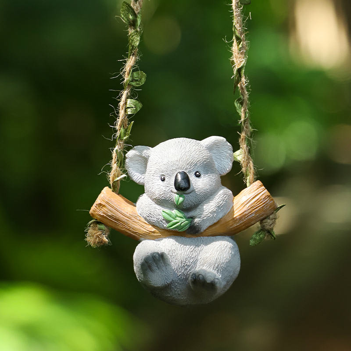 Cute grey koala figurine sitting on a wooden-style hanging swing holding green leaves, ideal for fairy garden accessories and miniature decor.