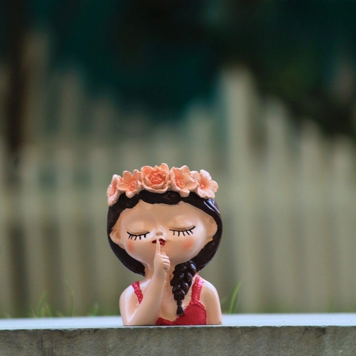 A miniature resin planter shaped like a girl’s head with a flower crown and her finger to her lips in a shushing gesture, set against a blurred garden background.