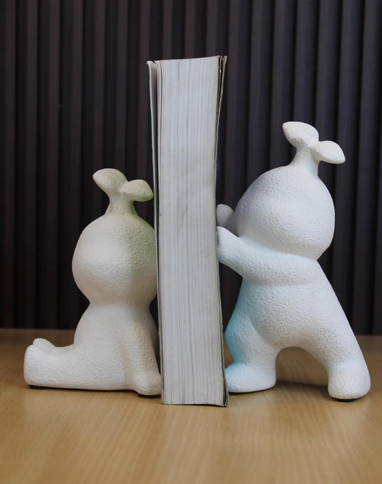 Two white, textured resin figurines shaped like stylized characters with small leaves on their heads, acting as bookends to hold a thick book upright on a wooden surface.