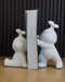 Two white, textured resin figurines shaped like stylized characters with small leaves on their heads, acting as bookends to hold a thick book upright on a wooden surface.