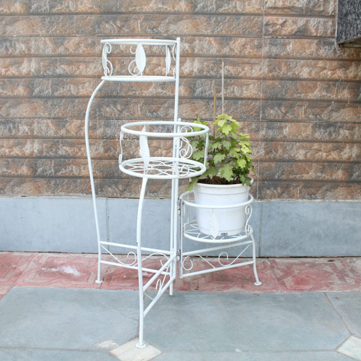 A 3-tier white metal plant stand with leaf patterns and scrollwork, holding a potted green plant against an outdoor stone wall.