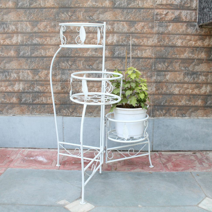 A 3-tier white metal plant stand with leaf patterns and scrollwork, holding a potted green plant against an outdoor stone wall.