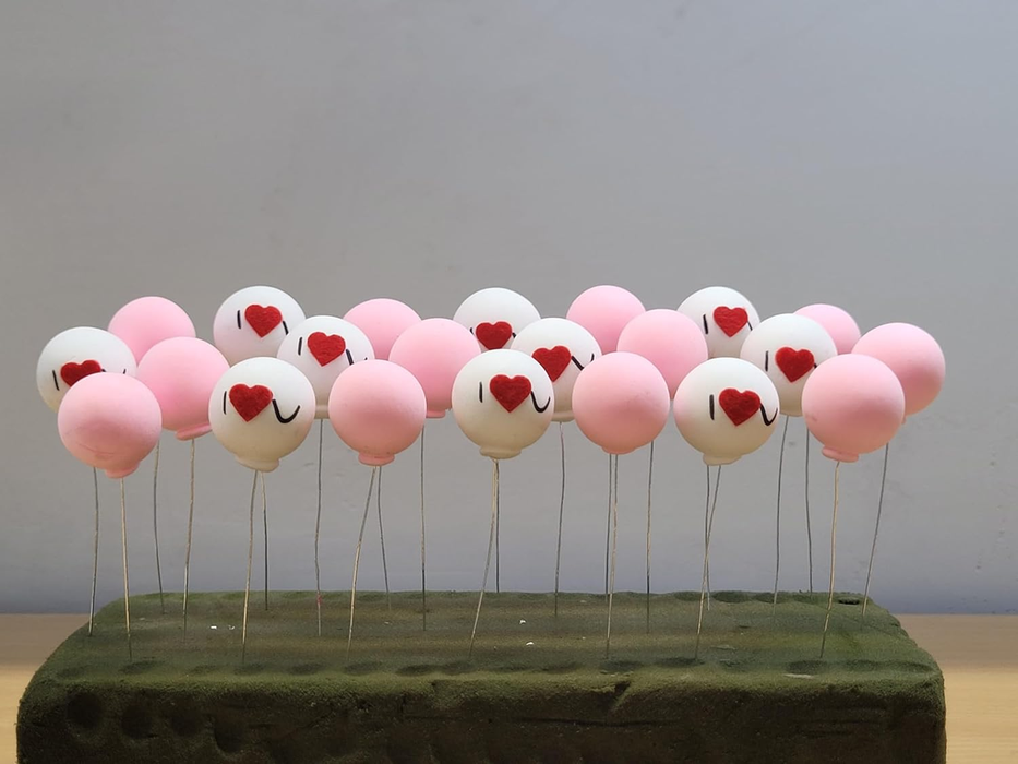 A row of miniature pink and white round balloon picks inserted into a green foam base. The white balloons feature red hearts and "I U" text, while the pink balloons are solid colors.