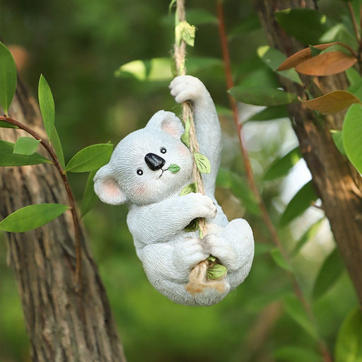 A charming hand-painted resin koala figurine hanging from a natural twine rope with green leaf accents, positioned between two tree trunks in a garden setting.