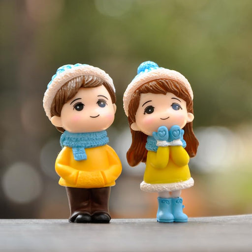Close-up of a pair of small resin miniature figurines, a boy and a girl, dressed in yellow winter jackets, blue hats, and scarves, positioned as a romantic couple showpiece.