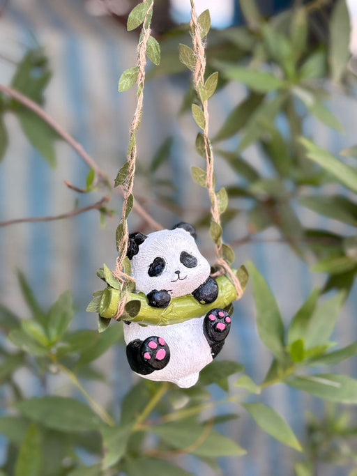 A small, decorative panda figurine hanging from a vine-wrapped rope swing. The panda is holding a green bamboo branch and has pink paw pads, set against a blurred background of green garden leaves.